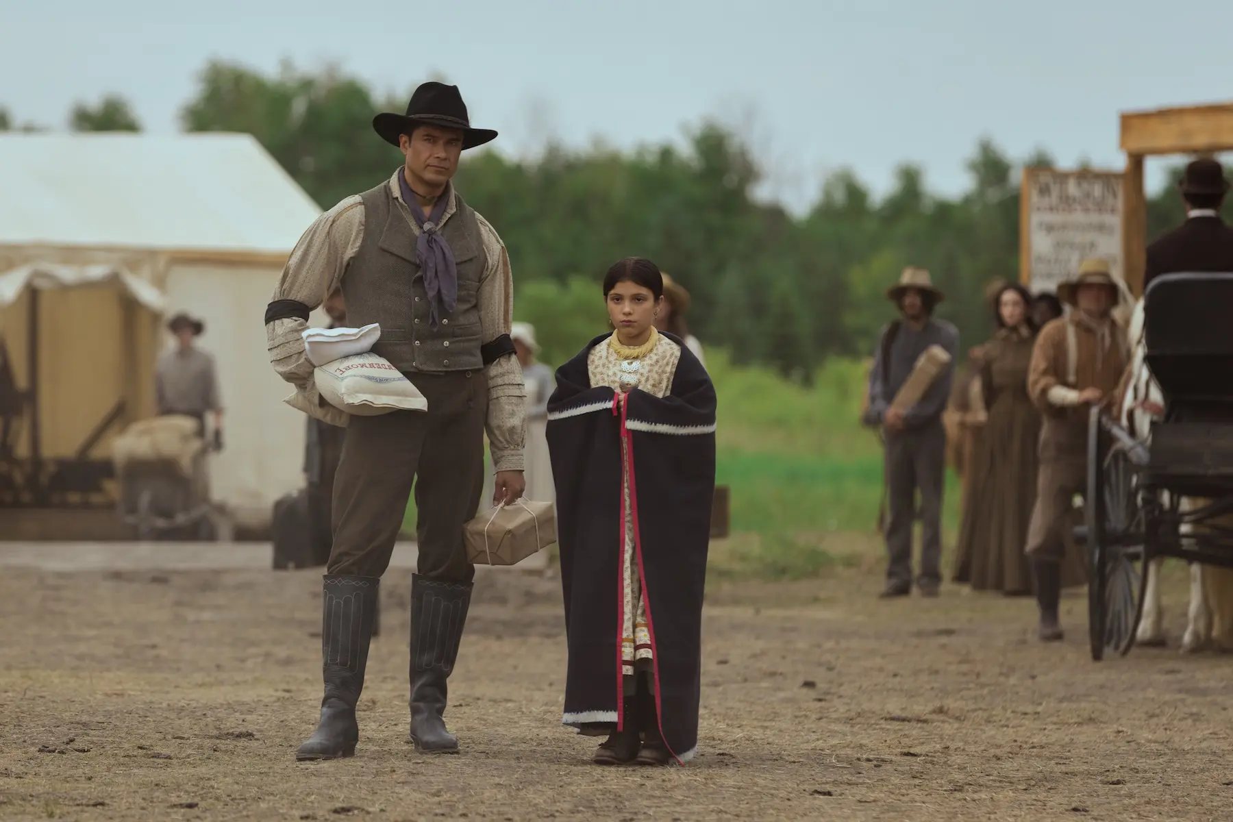 A man in a wide-brimmed hat carries a flour sack and a paper-wrapped parcel as he walks beside a girl wrapped in a dark wool blanket with a red stripe, along a dirt street in a frontier town.