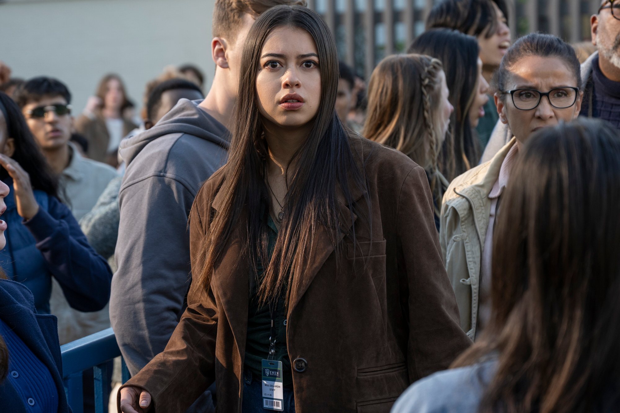 Amber Midthunder, in a brown coat and a staff identification badge, stands among a crowd outdoors in a scene from Carrie.