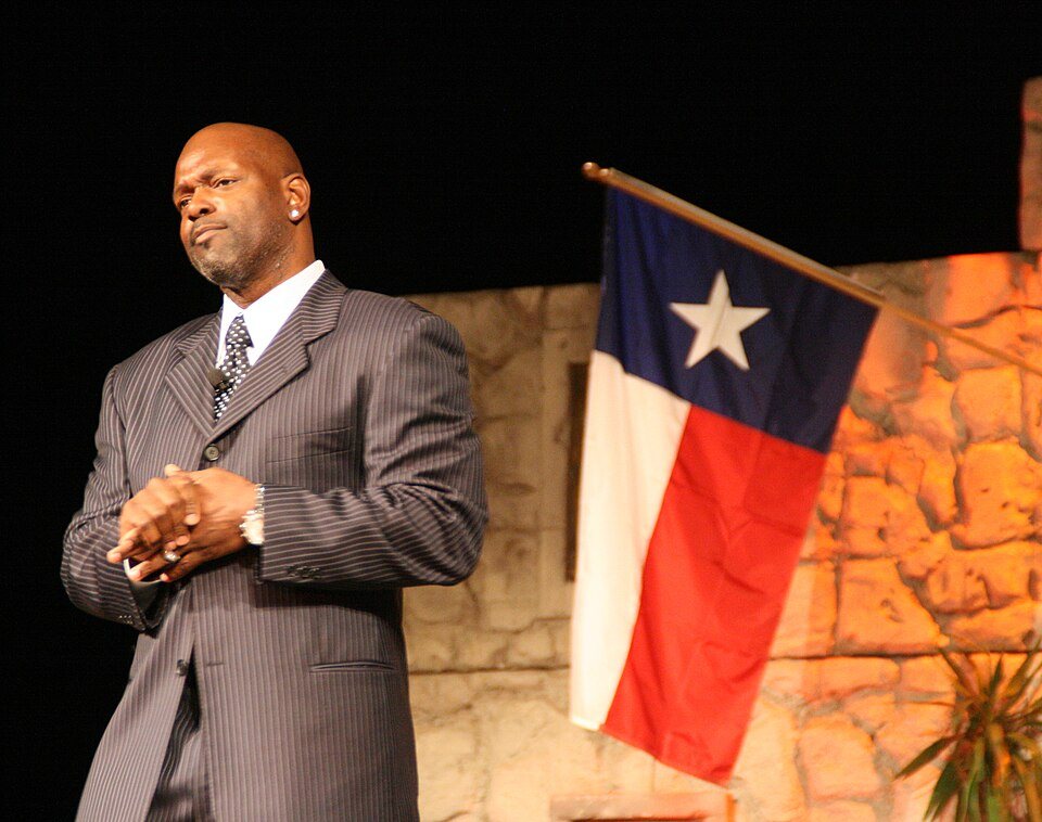 Emmitt Smith, in a pinstriped suit, stands on a stage beside a Texas flag.