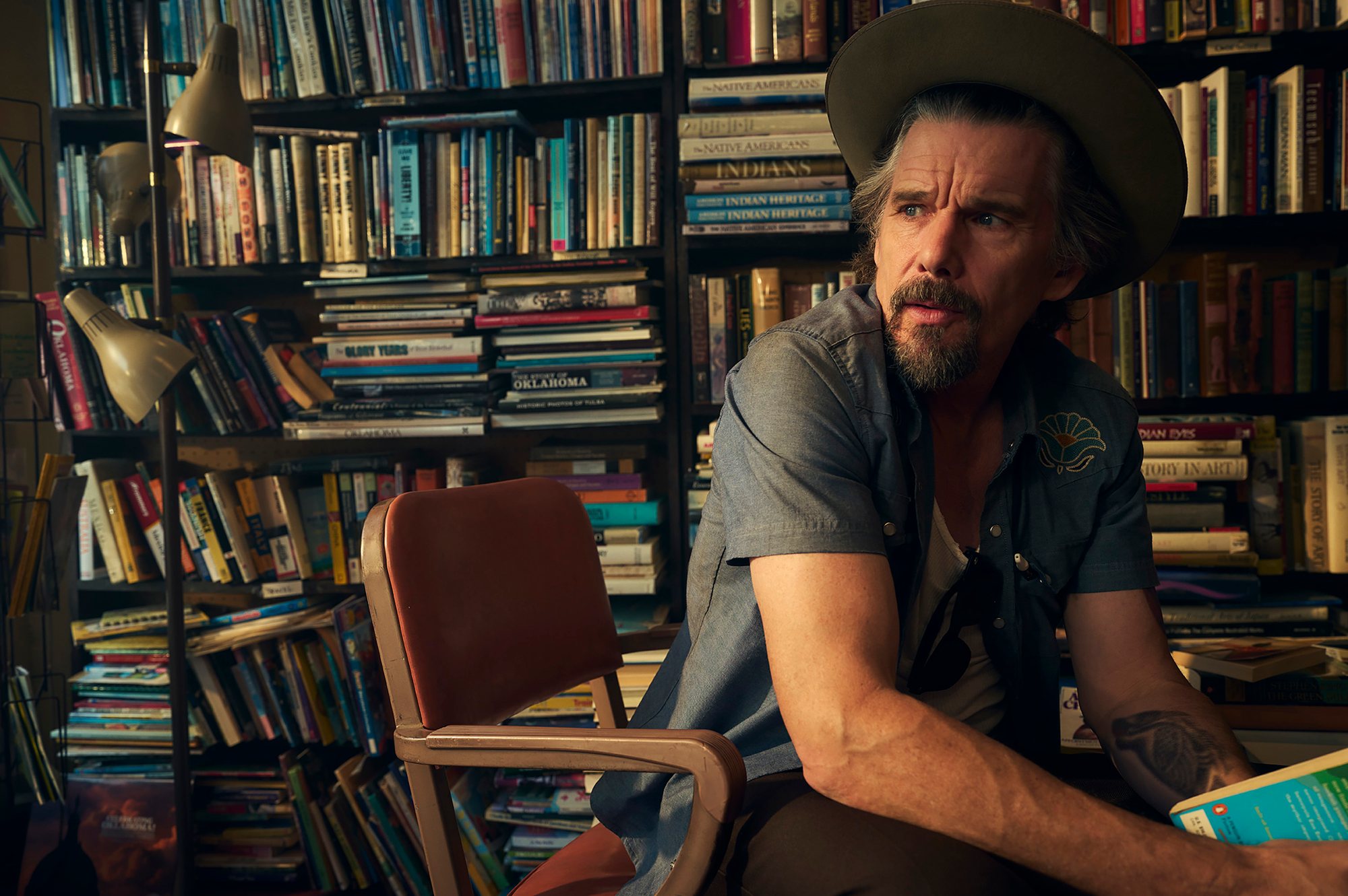 Ethan Hawke, in a wide-brimmed hat and denim shirt, sits in a chair in front of crowded bookshelves.