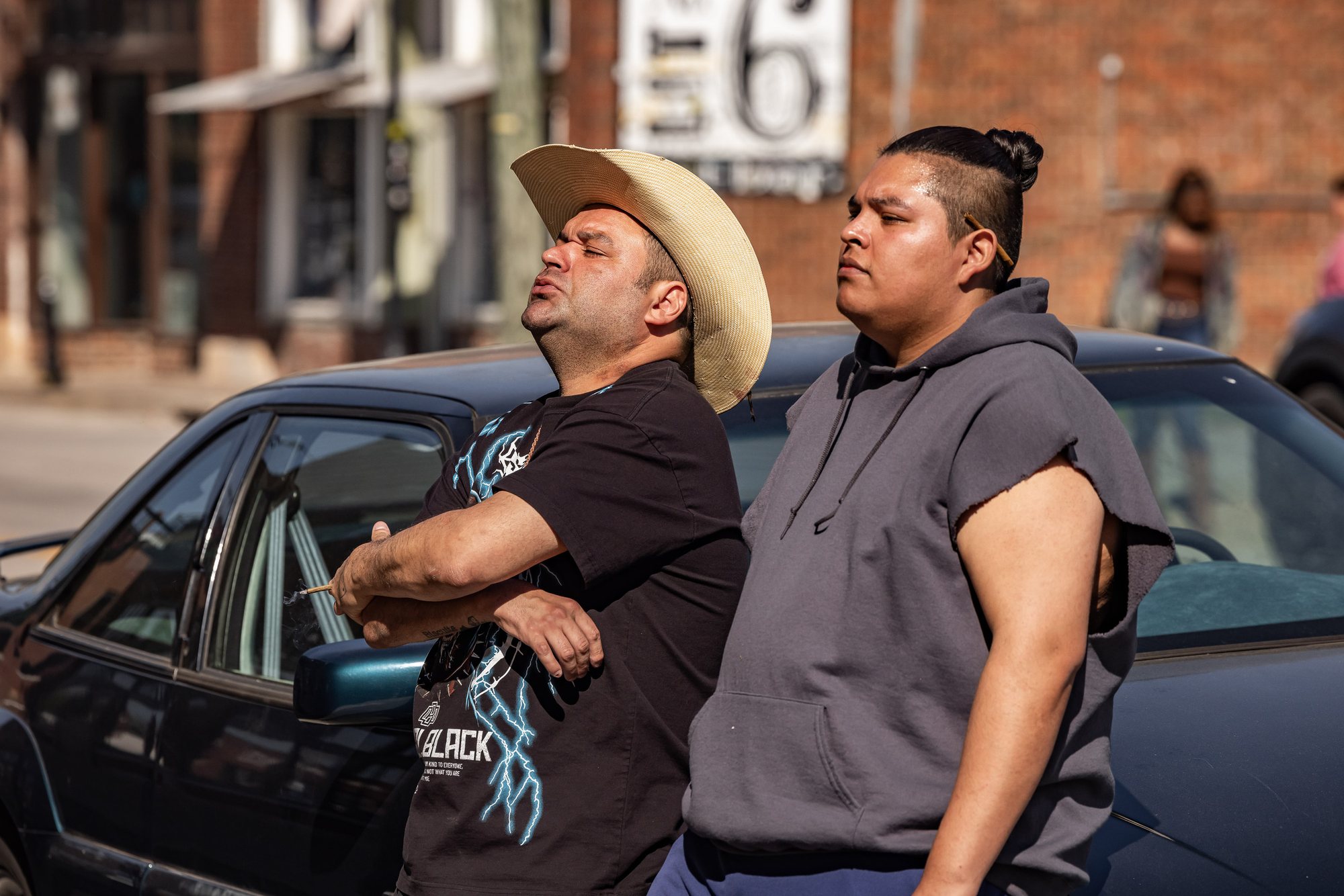 Cody Lightning, in a straw cowboy hat, and Jude Barnett, in a sleeveless grey hoodie, lean against a dark car on a city street.