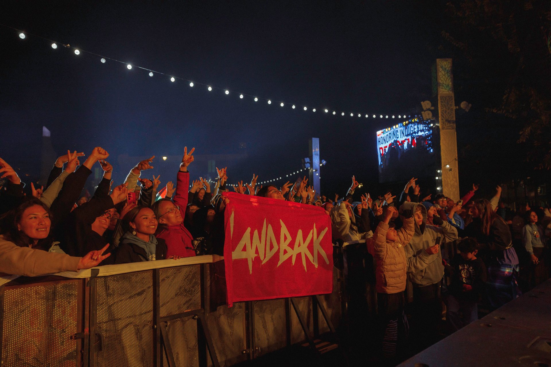 Concertgoers at the barricade raise their hands beside a red LANDBACK banner, under strings of lights.