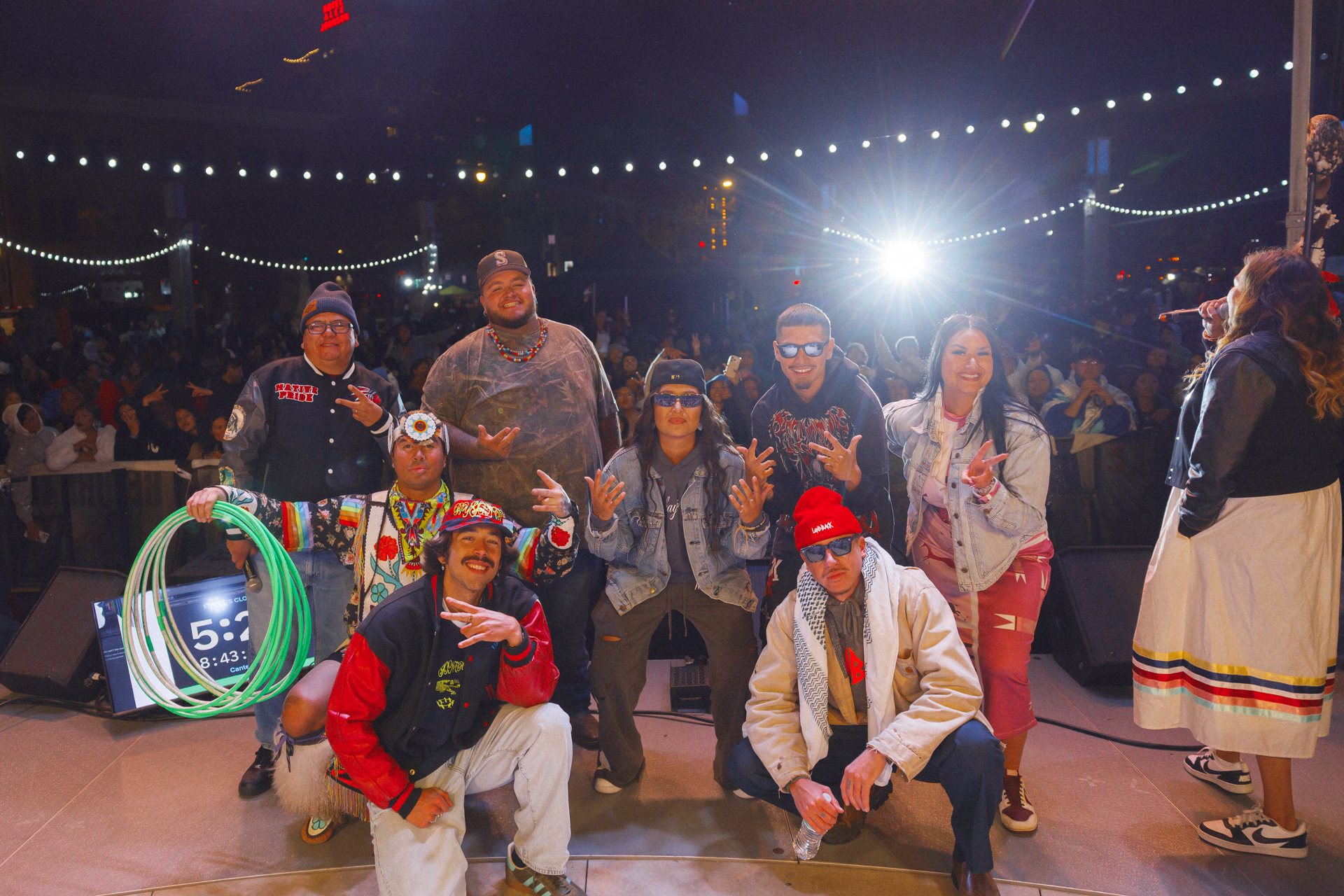 Eight performers pose together in two rows on stage, with Macklemore at the front right.