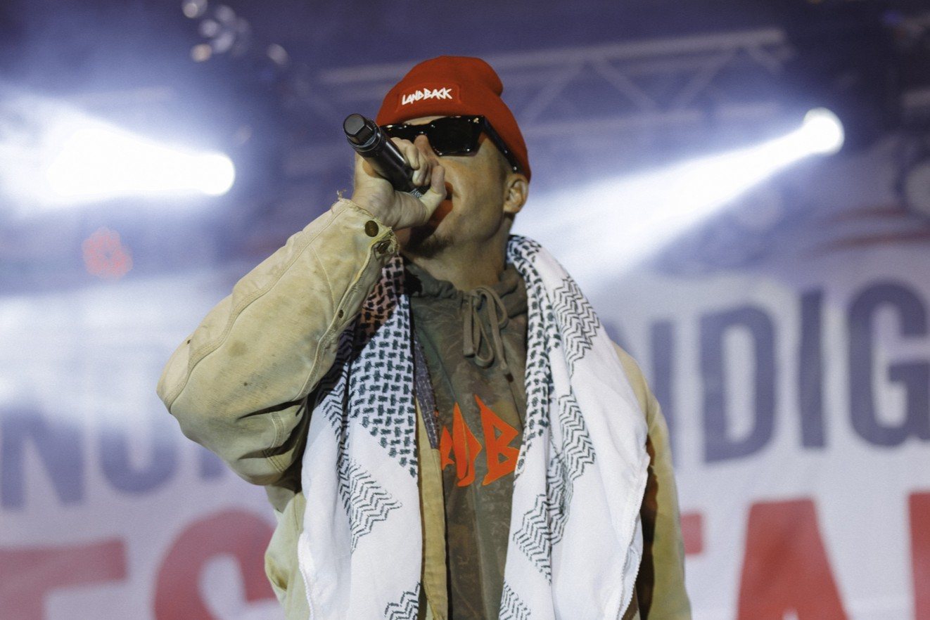 Macklemore, in a red LANDBACK beanie, sunglasses and a keffiyeh, raps into a microphone on stage.