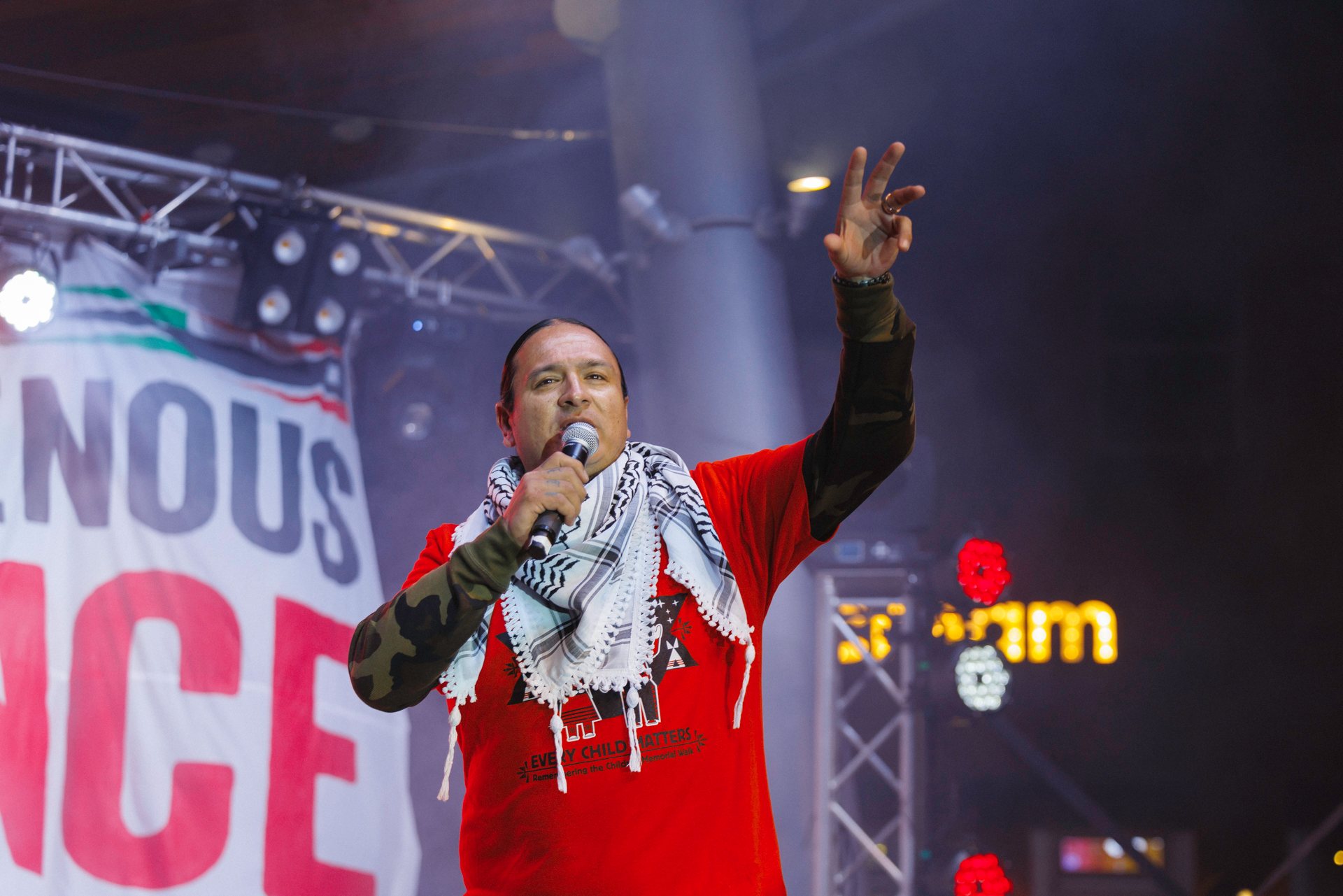 Nick Tilsen, in a red Every Child Matters shirt and a keffiyeh, speaks into a microphone with one hand raised.