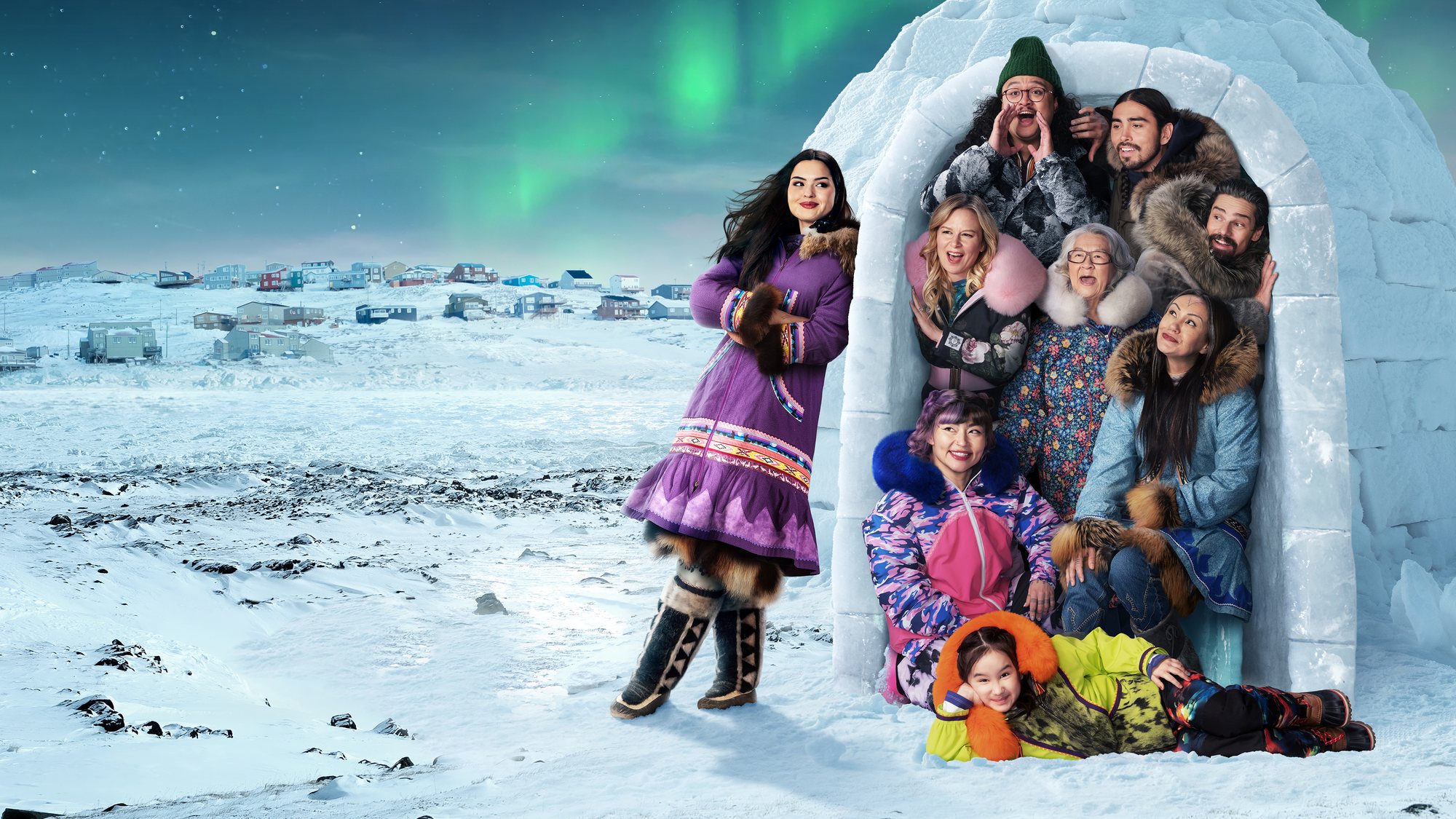The North of North cast poses in and beside an igloo on snowy land outside a hillside town under green northern lights, with Anna Lambe standing at left in a purple parka.