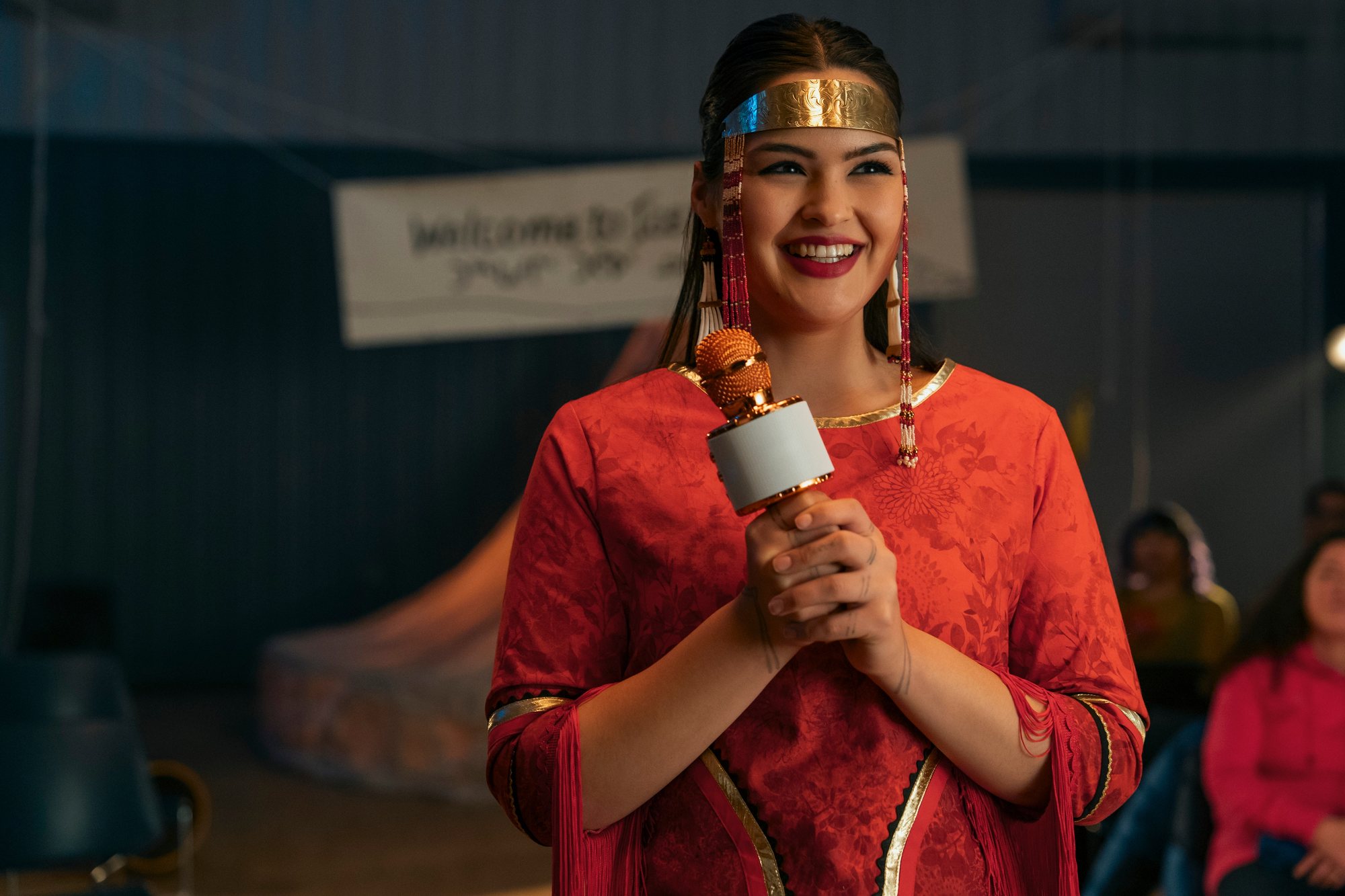 Anna Lambe as Siaja smiles while holding a microphone in a community hall, wearing a red dress, a gold headband and long beaded earrings.