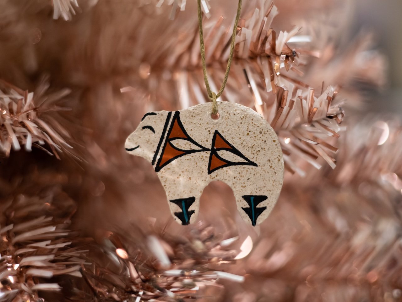 A speckled white ceramic bear ornament painted with black, orange and turquoise designs hangs from a twine loop on a rose-gold artificial tree.