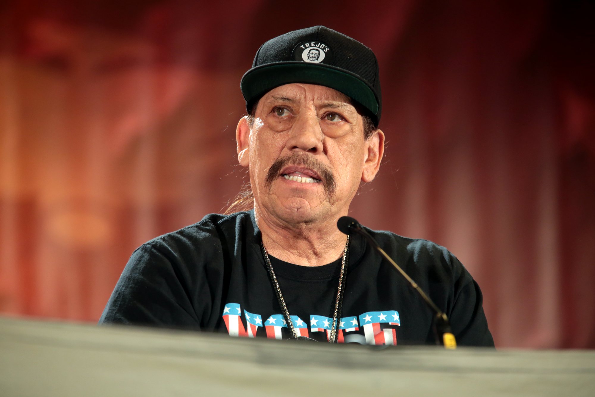 Danny Trejo, in a black cap and T-shirt, speaks into a microphone at a convention panel.