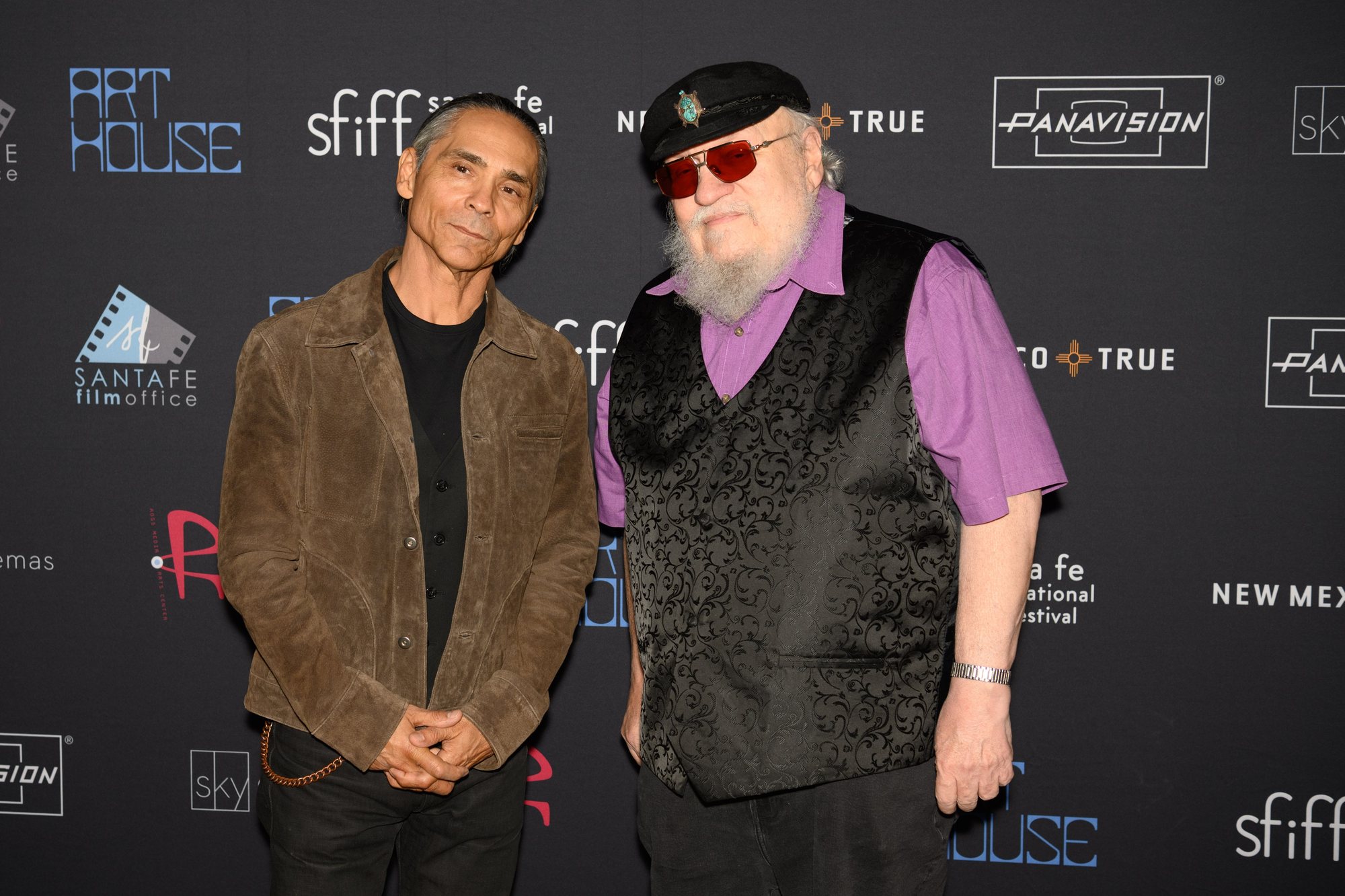 Zahn McClarnon, in a brown suede jacket, stands beside George R.R. Martin in front of a Santa Fe International Film Festival backdrop.