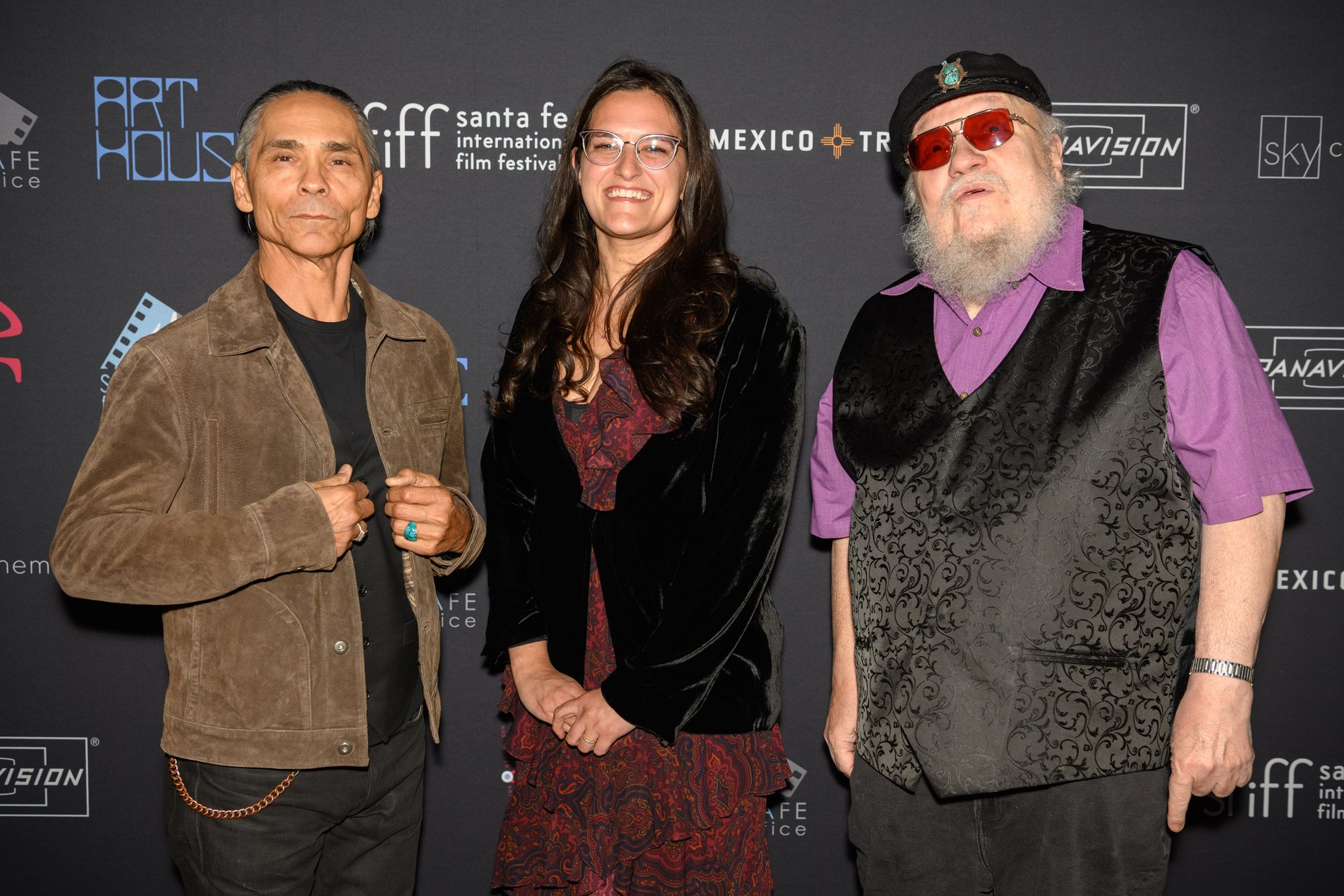 Zahn McClarnon, festival executive director Liesette Bailey and George R.R. Martin stand together in front of a Santa Fe International Film Festival backdrop.
